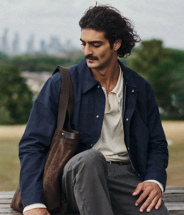 Man sitting outdoors wearing a NN.07 navy jacket with a brown bag, looking away from the camera.7